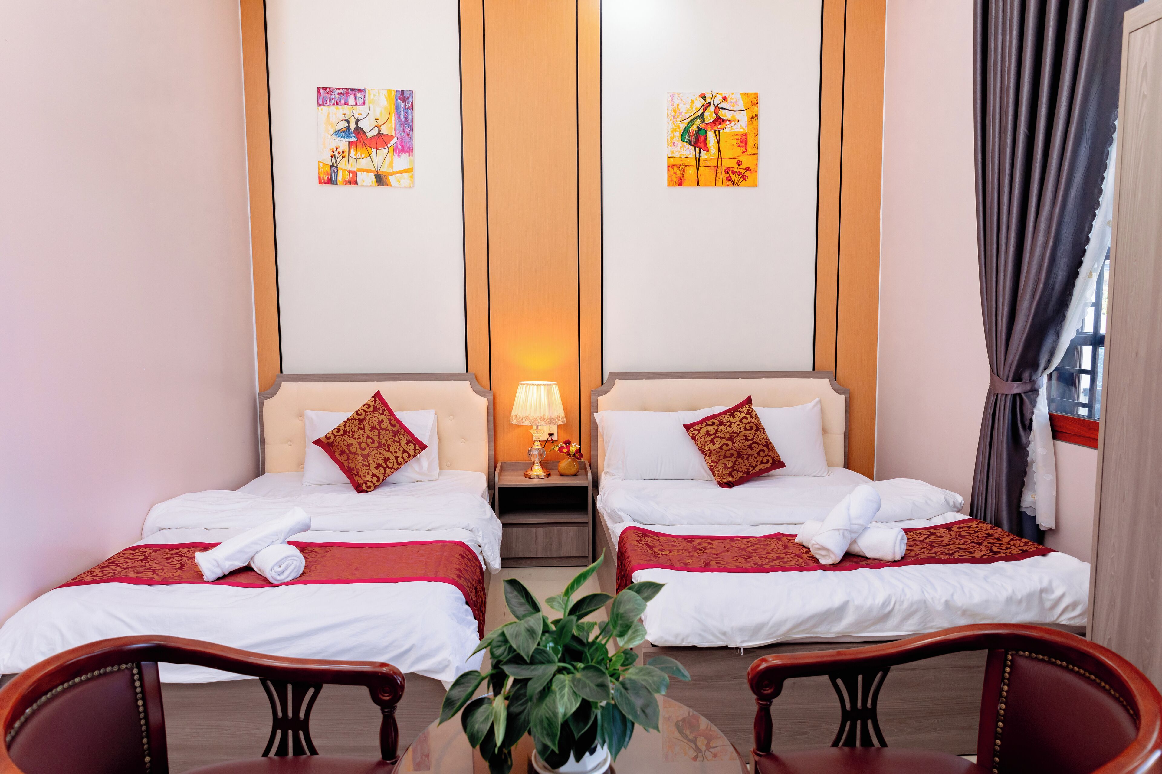 A hotel room with two beds, a table with chairs, a plant, and artwork on the walls.