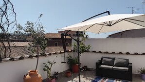 Eine Dachterrasse mit weißem Sonnenschirm, Topfpflanzen, einer schwarzen Couch und einem gemusterten Teppich.