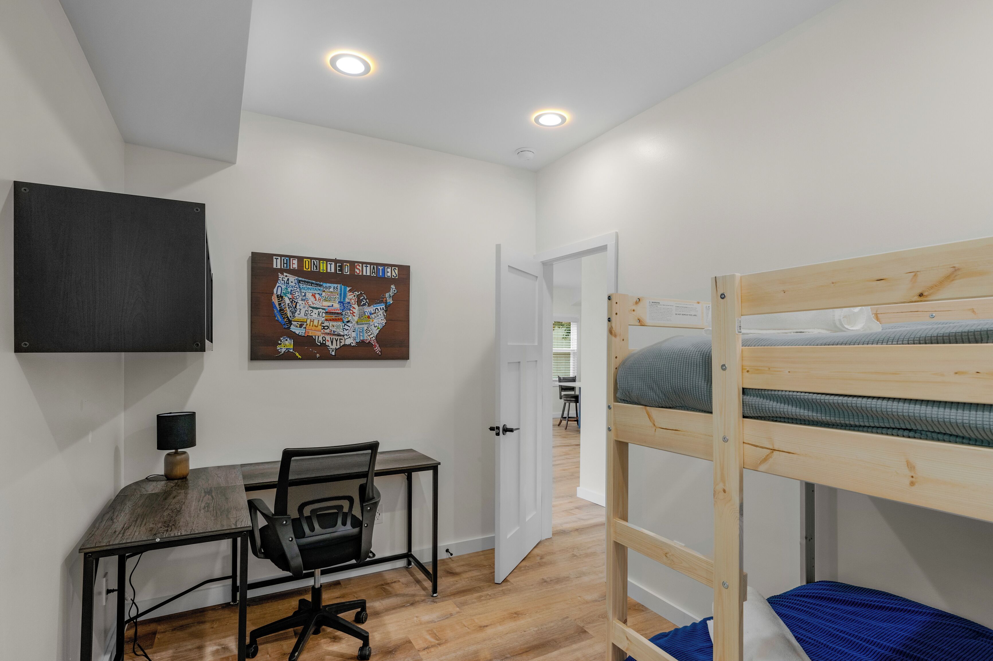 Une chambre avec un lit superposé, un bureau avec une chaise, une carte accrochée au mur et une porte donnant sur une autre pièce.