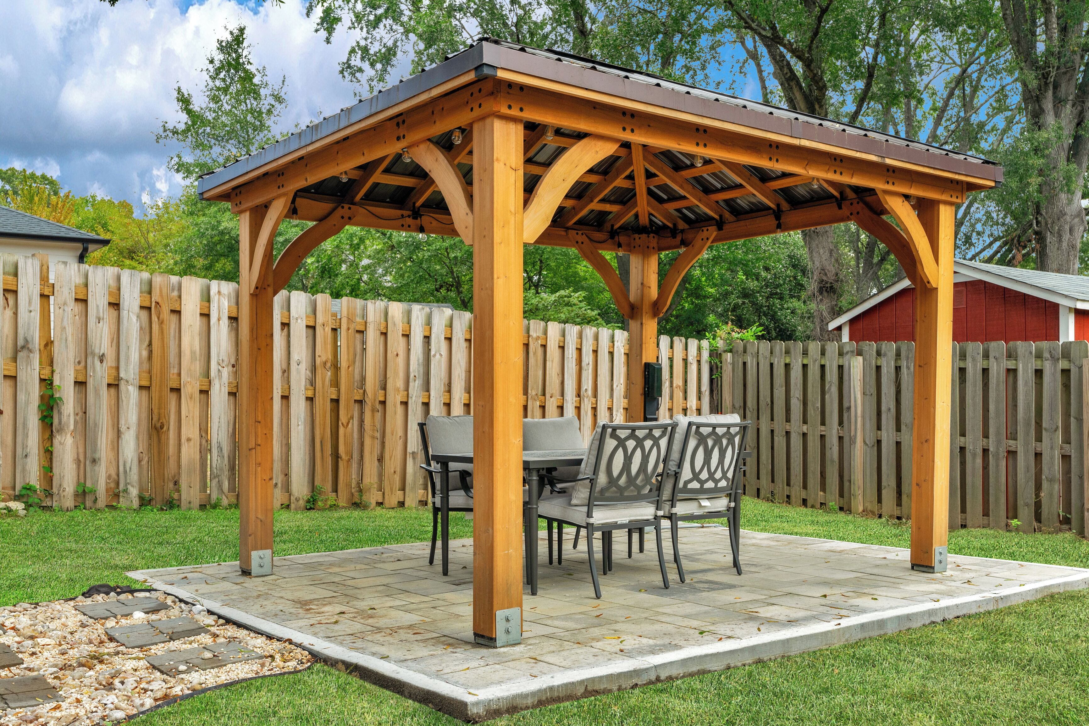 A wooden pergola with a table and chairs underneath, surrounded by a wooden fence and a grassy area.
