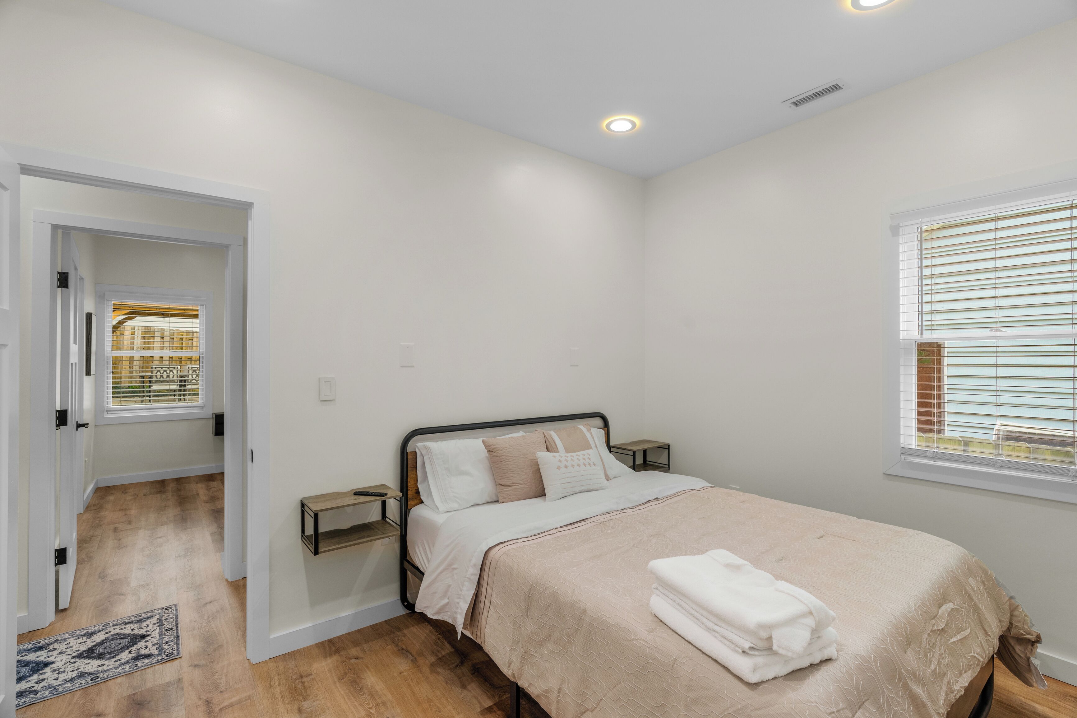 A bedroom with a bed, bedside tables, a window with blinds, a door leading to another room, and a wooden floor.