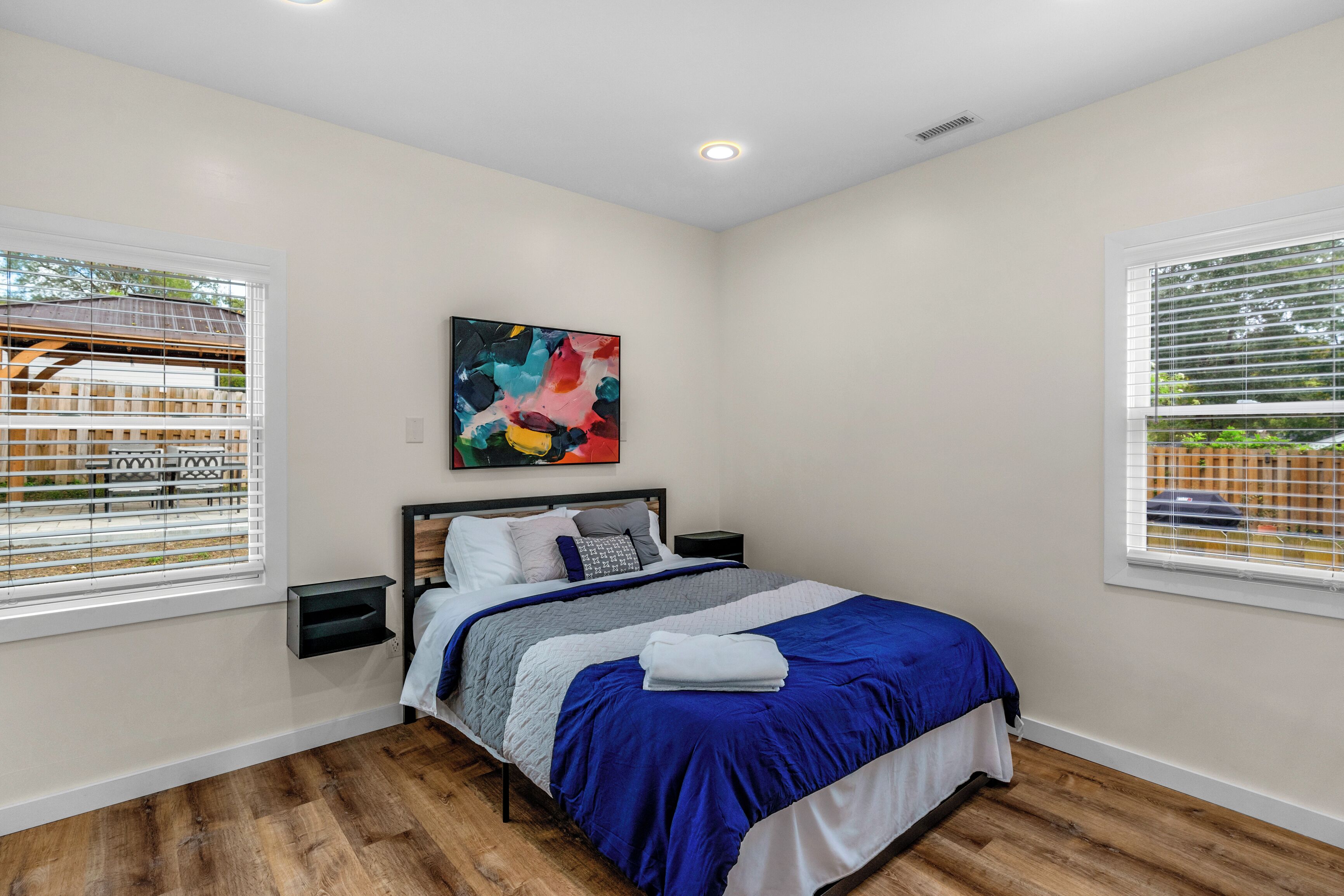 A bedroom with a bed, a nightstand, a wall-mounted painting, and a window with blinds.