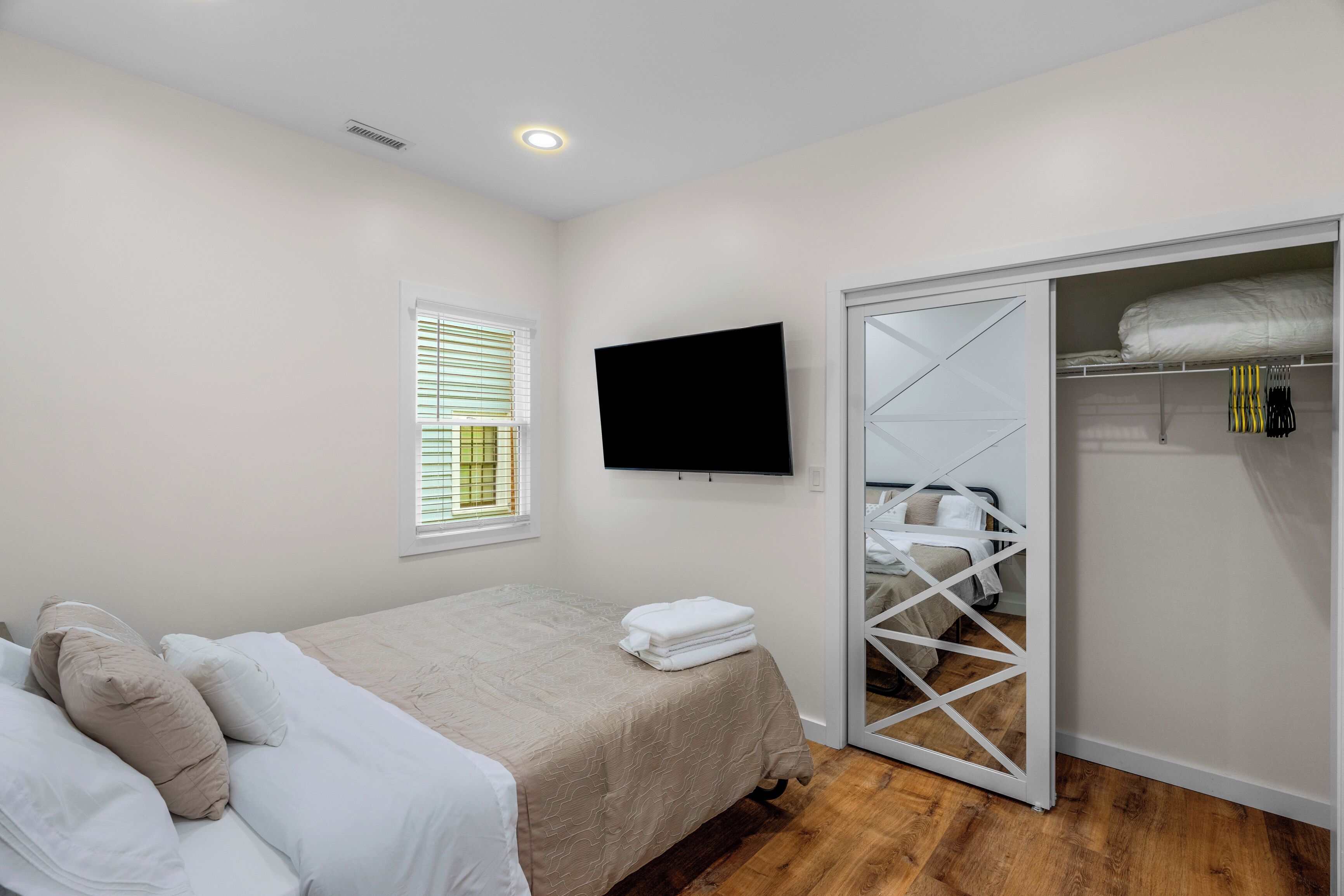 Une chambre à coucher comprenant un lit, un téléviseur fixé au mur, une armoire avec une porte miroir et une fenêtre avec des stores.