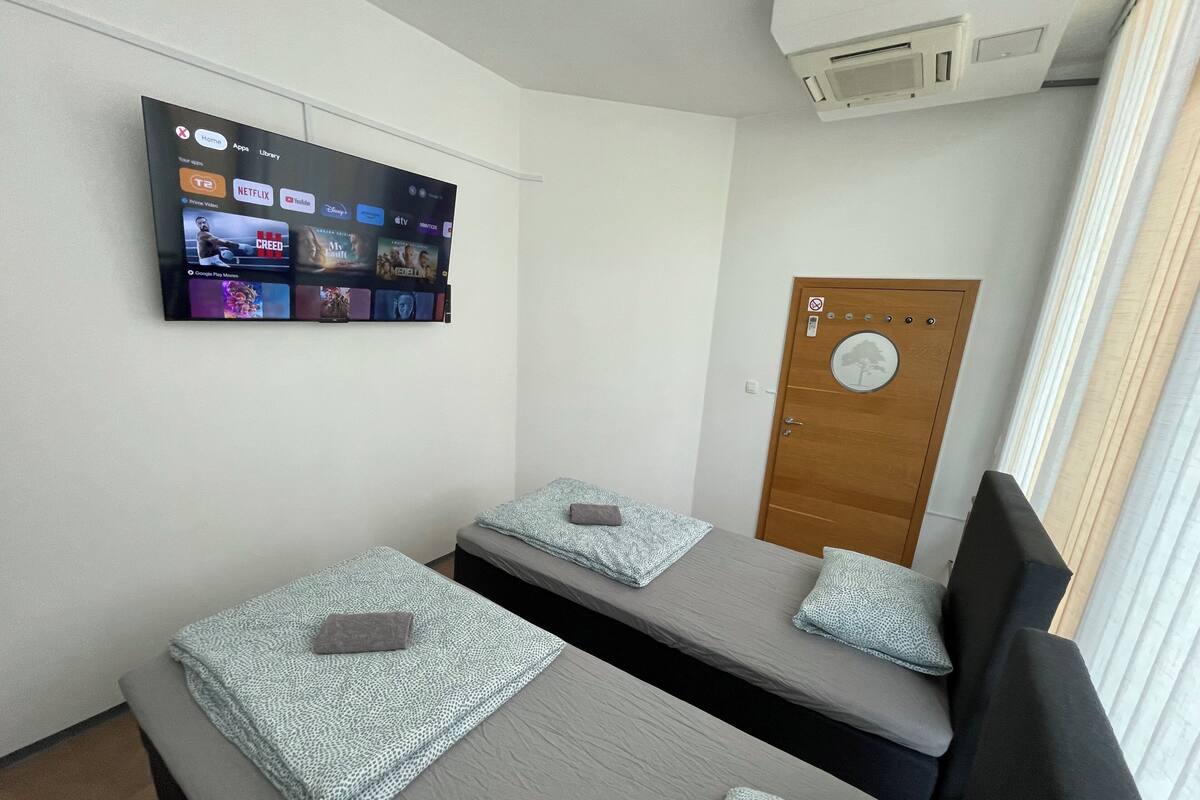 Habitación de hotel con dos camas, un televisor montado que muestra varios servicios de streaming, una puerta de madera con un adhesivo de árbol y un equipo de aire acondicionado.