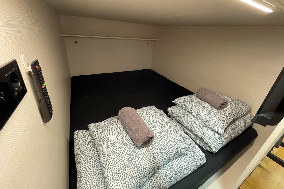 Una habitación pequeña con una cama individual, un colchón negro y almohadas estampadas, un control remoto y una lámpara fijada a la pared.