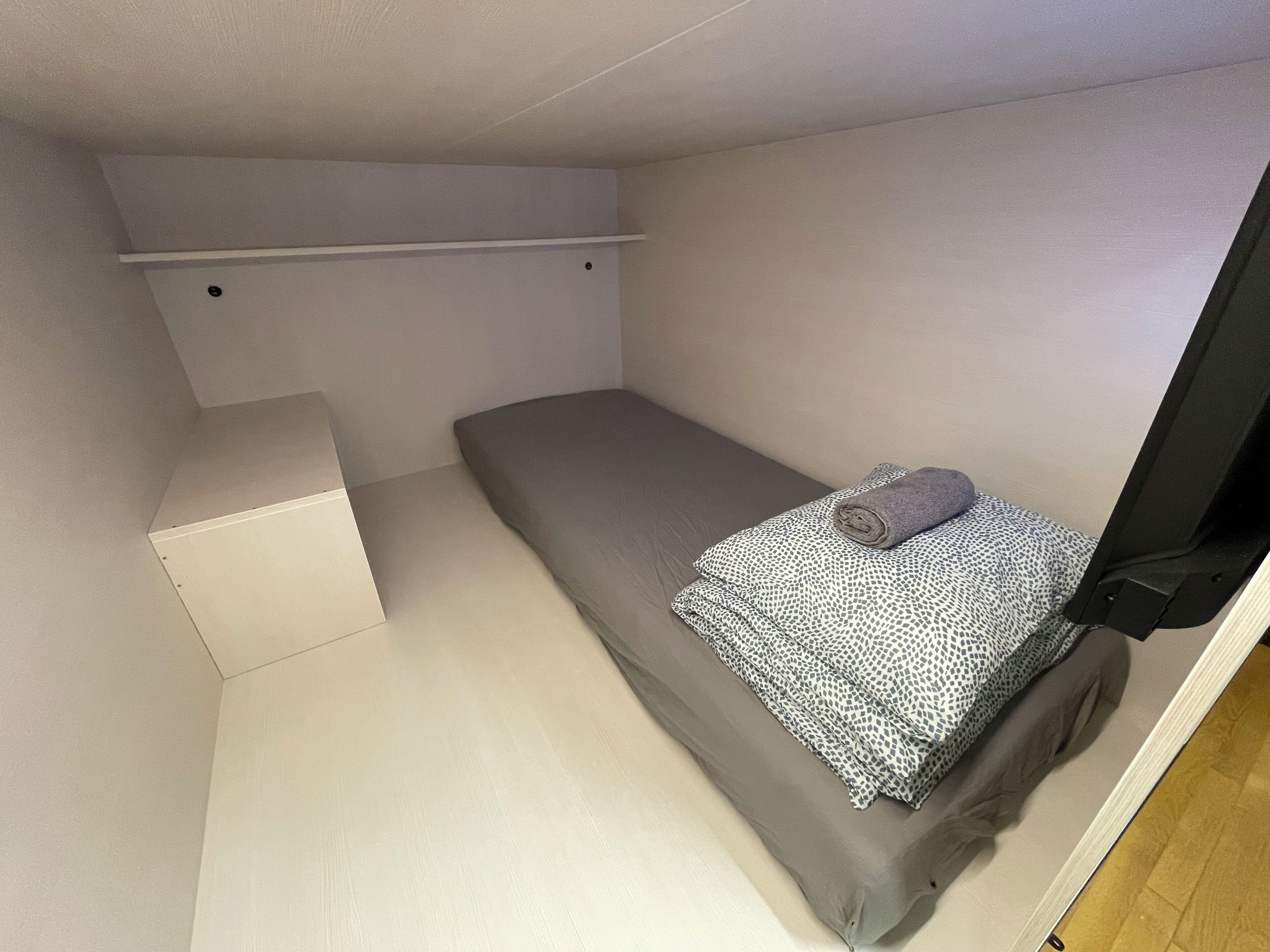 A small, single-bed room with a grey mattress, a patterned blanket, and a rolled-up towel. There is a white shelf above the bed and a wooden floor.