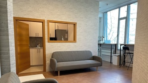 A modern living room with a grey sofa, a wooden door leading to a kitchen, and a large window providing natural light. - Sleep&Go Hostel (Ljubljana)