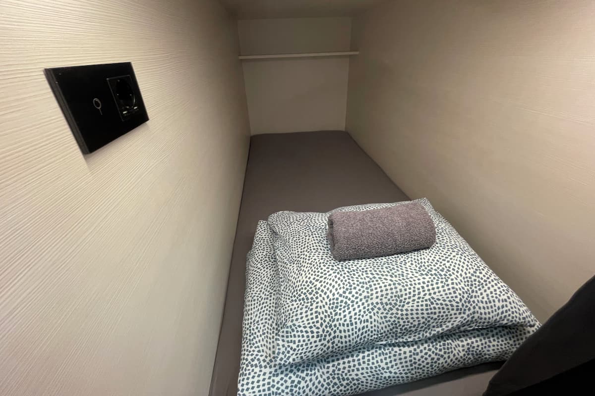 Una habitación pequeña y estrecha con una cama individual, un panel de control en la pared y un techo sencillo.