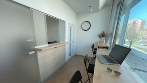 A modern office with a desk, chairs, a laptop, a clock, and a window with a view of buildings and trees. - Sleep&Go Hostel (Ljubljana)