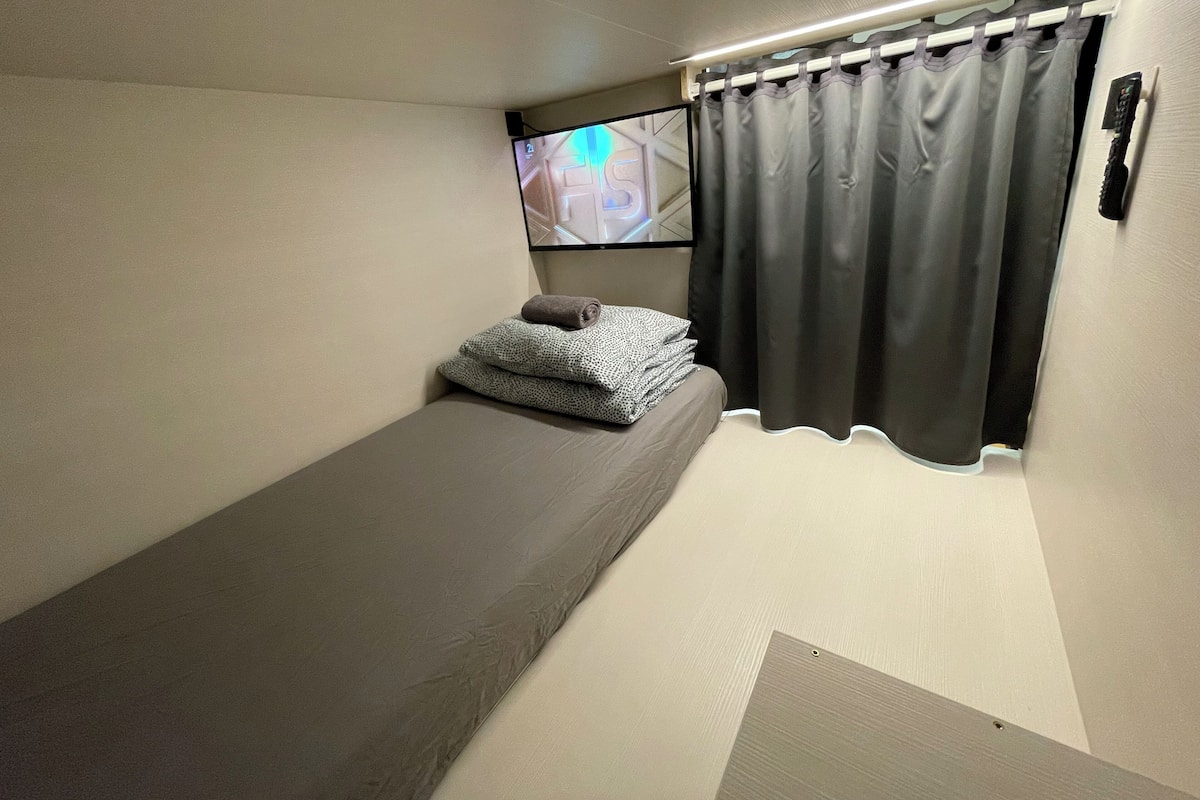 Una habitación pequeña con cama individual, ventana, cortina y televisor montado en la pared.