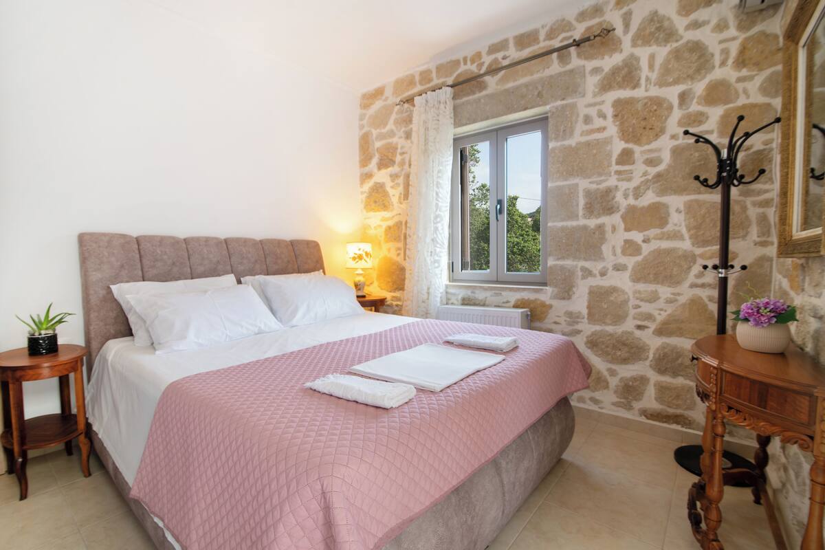Un dormitorio con pared de piedra, cama con colcha rosa, mesita de noche con lámpara y ventana con cortinas.