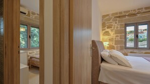 Une chambre avec un mur en pierre, un lit recouvert de draps blancs et une fenêtre donnant sur des immeubles.