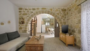 Un salon avec un mur en pierre, un canapé, une table basse en bois, un téléviseur posé sur un meuble en bois et un escalier menant à la cuisine.