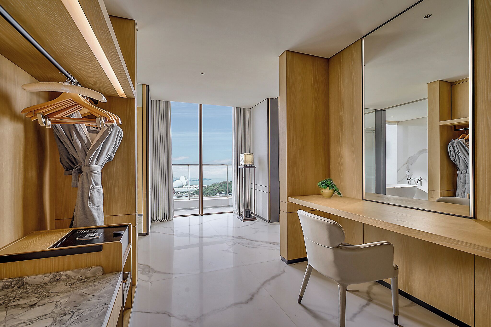Una habitación de hotel moderna con un ventanal grande, un escritorio con una silla, un tocador con espejo y un perchero.