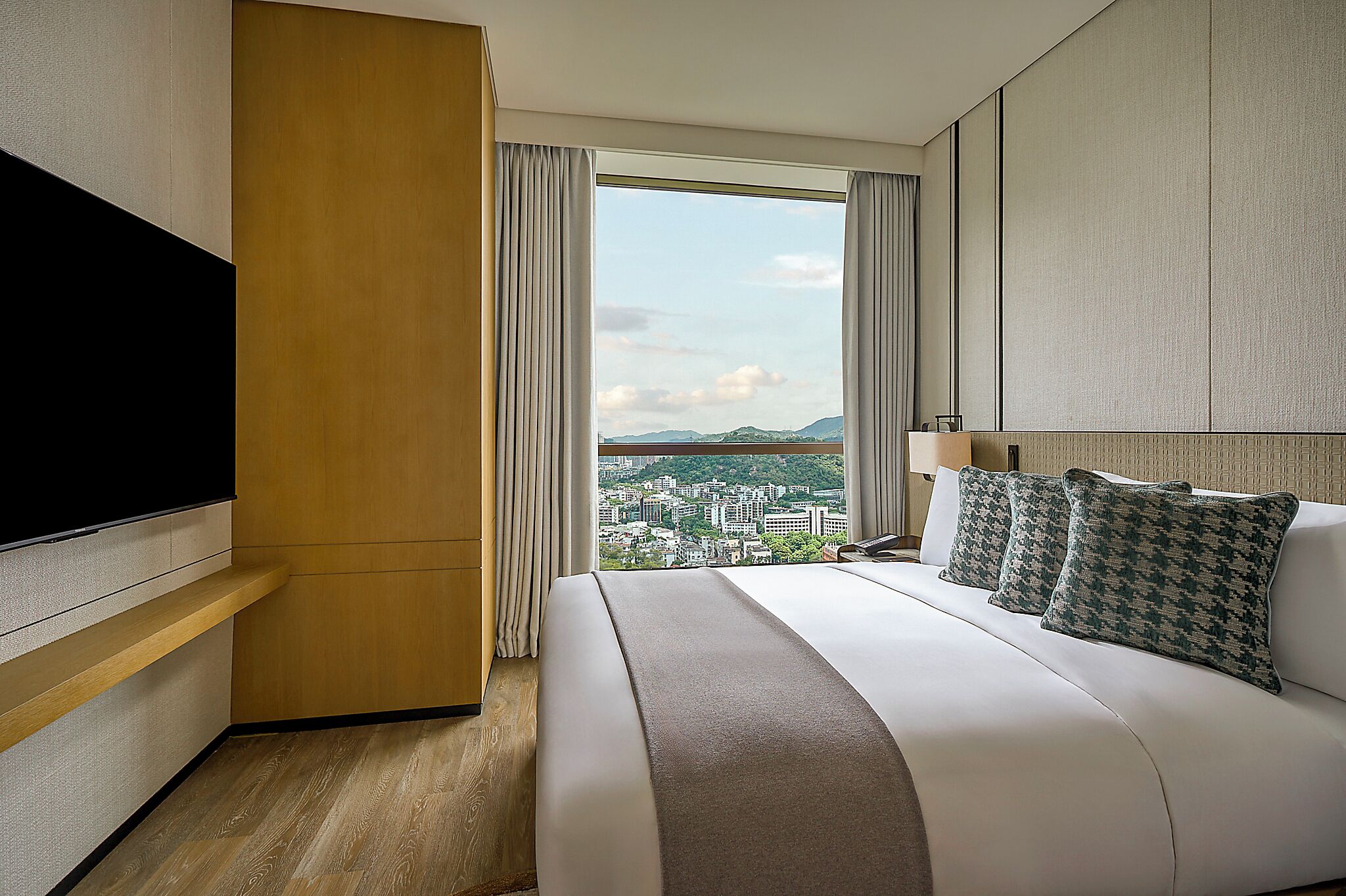 A hotel room with a bed, a TV, a window with curtains, and a view of a cityscape.