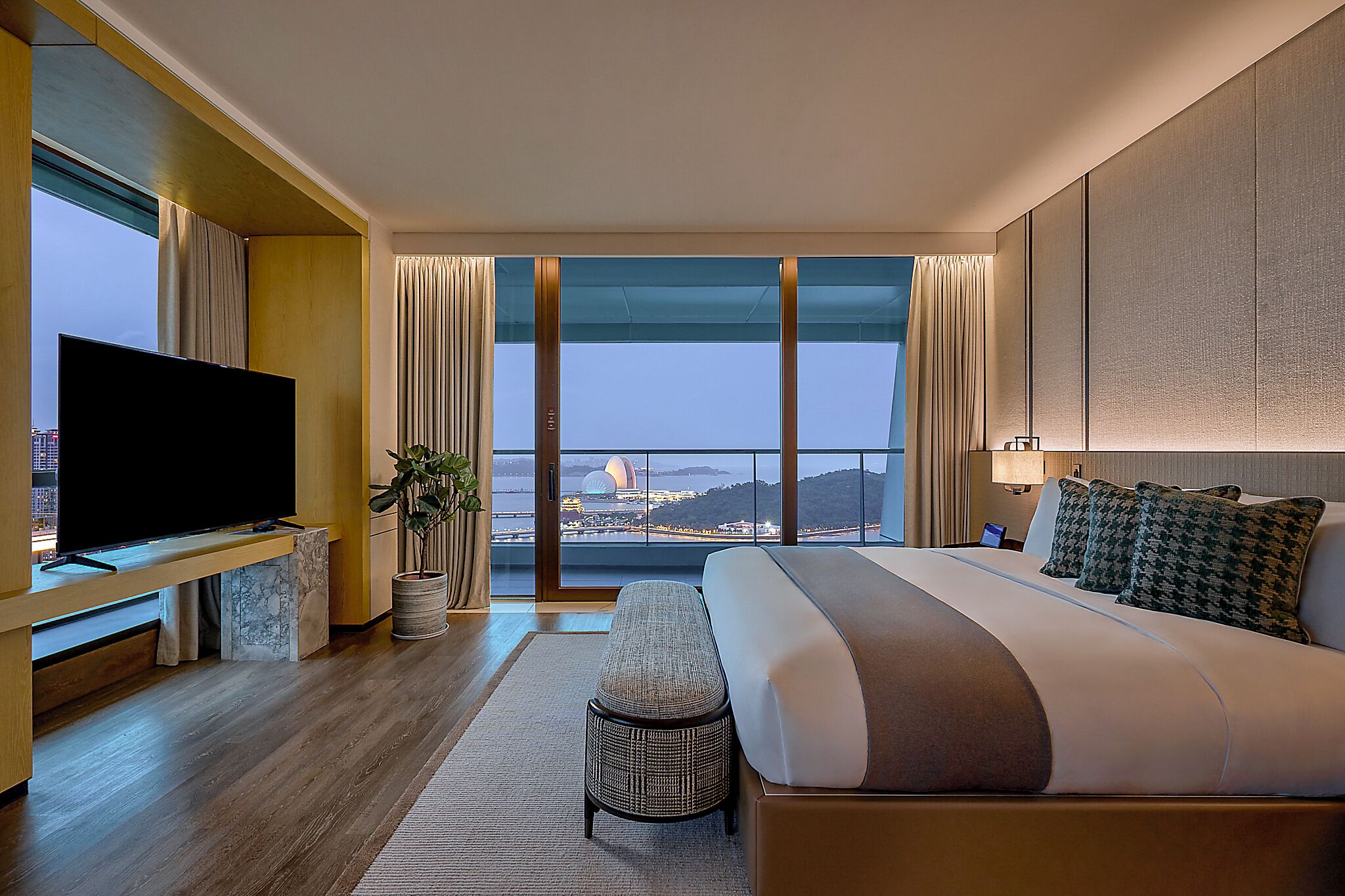 A modern hotel room with a large bed, a flat-screen TV, a view of the cityscape, and a balcony.
