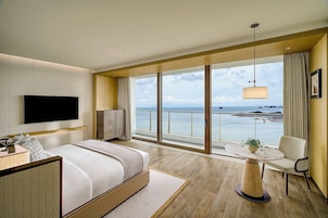 Quarto de hotel moderno com uma cama grande, televisão, varanda com vista para o mar e uma área de refeições com mesa e cadeira.