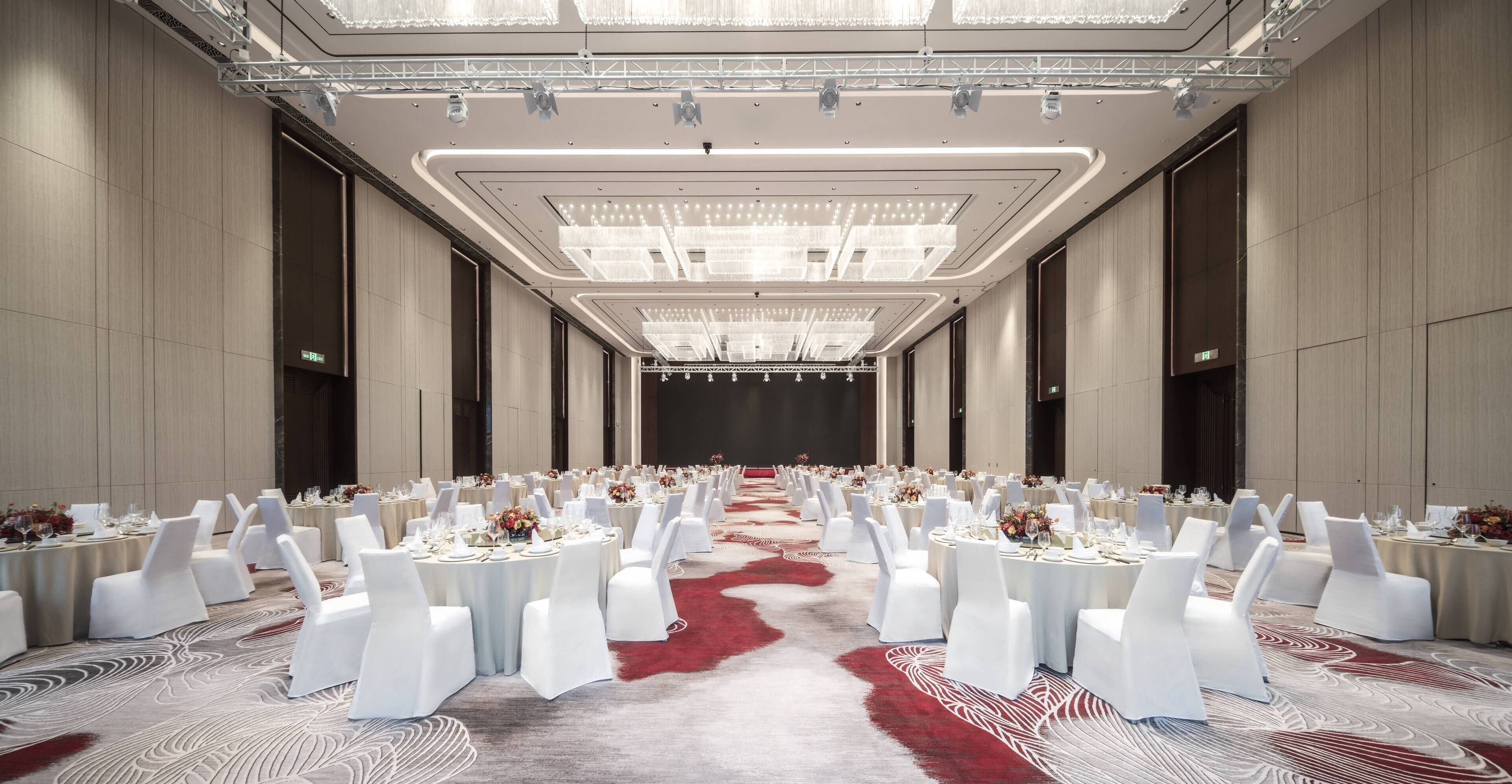 Un amplio salón de banquetes con filas de mesas redondas, sillas blancas y una alfombra roja.