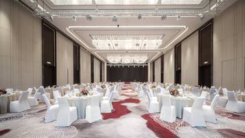Un amplio salón de banquetes con filas de mesas redondas, sillas blancas y una alfombra roja.