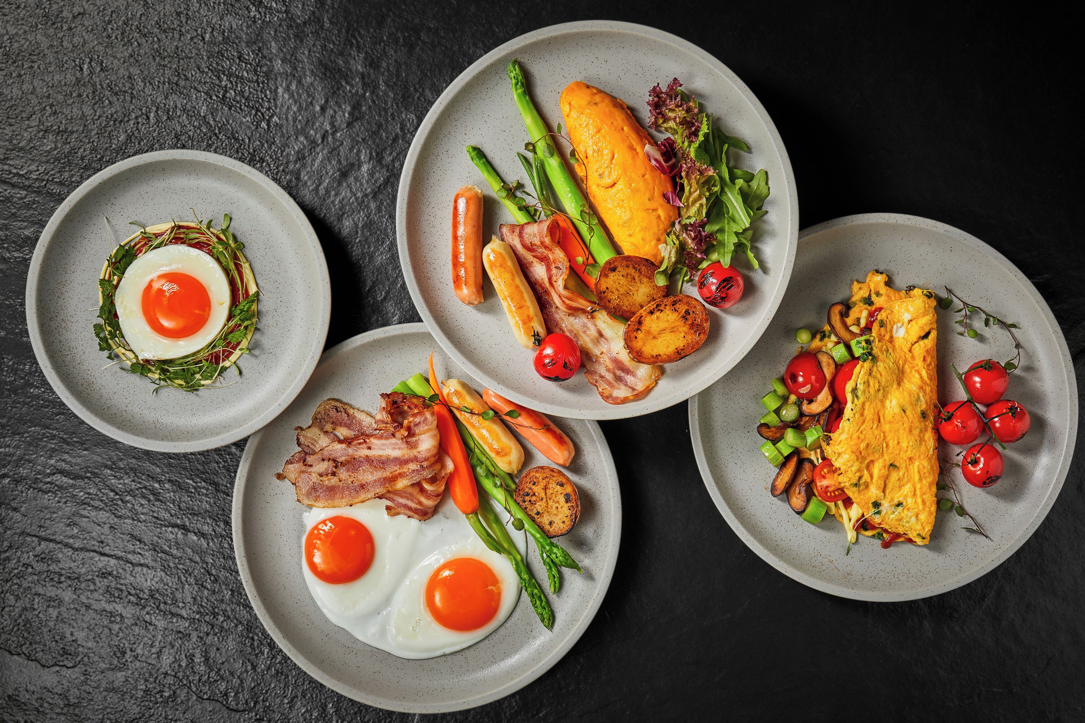 Verschiedene Gerichte mit Eiern, Spargel, Tomaten und Speck auf Tellern.
