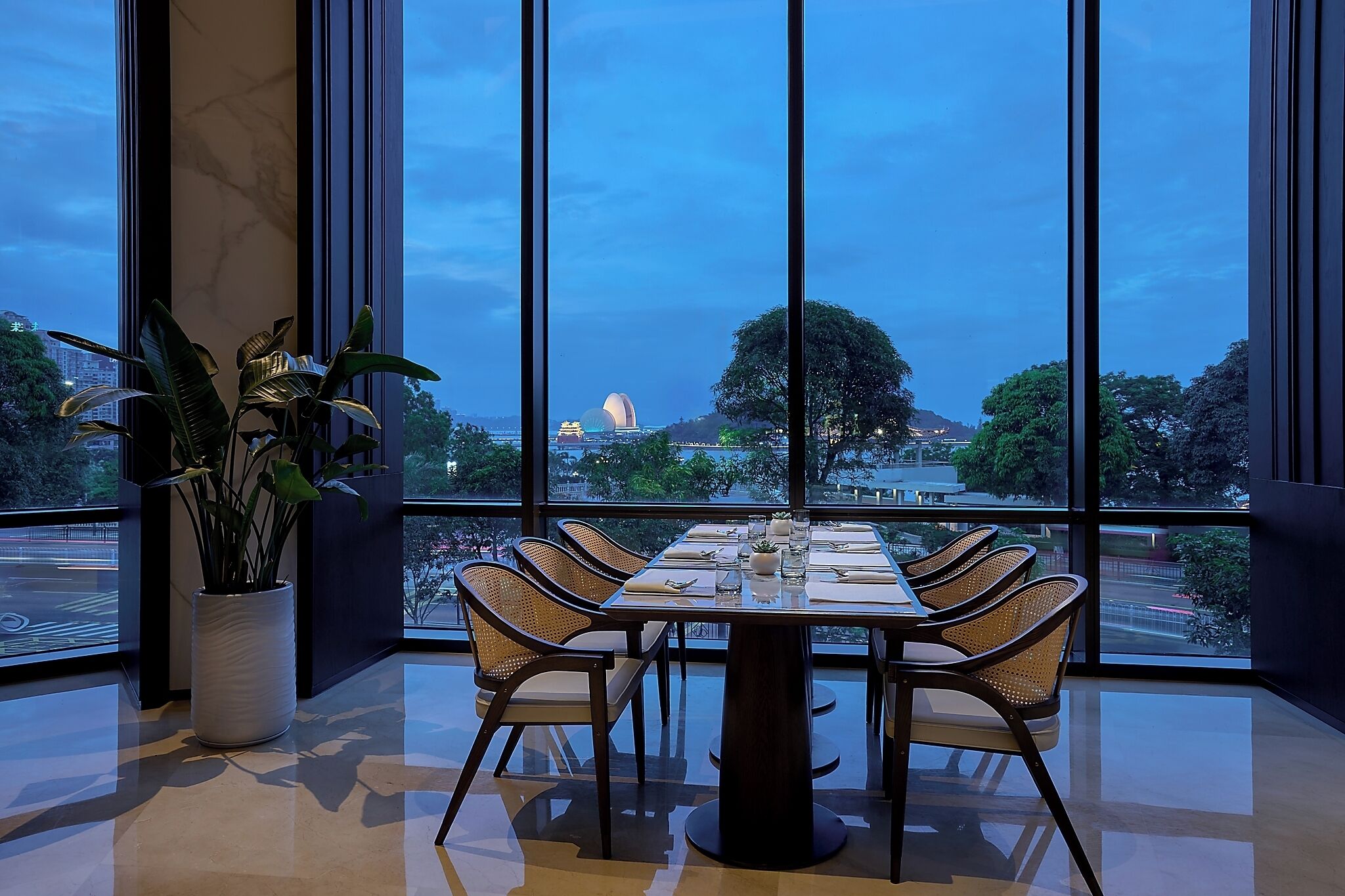 A dining area with a table set for six, large windows, and a potted plant.