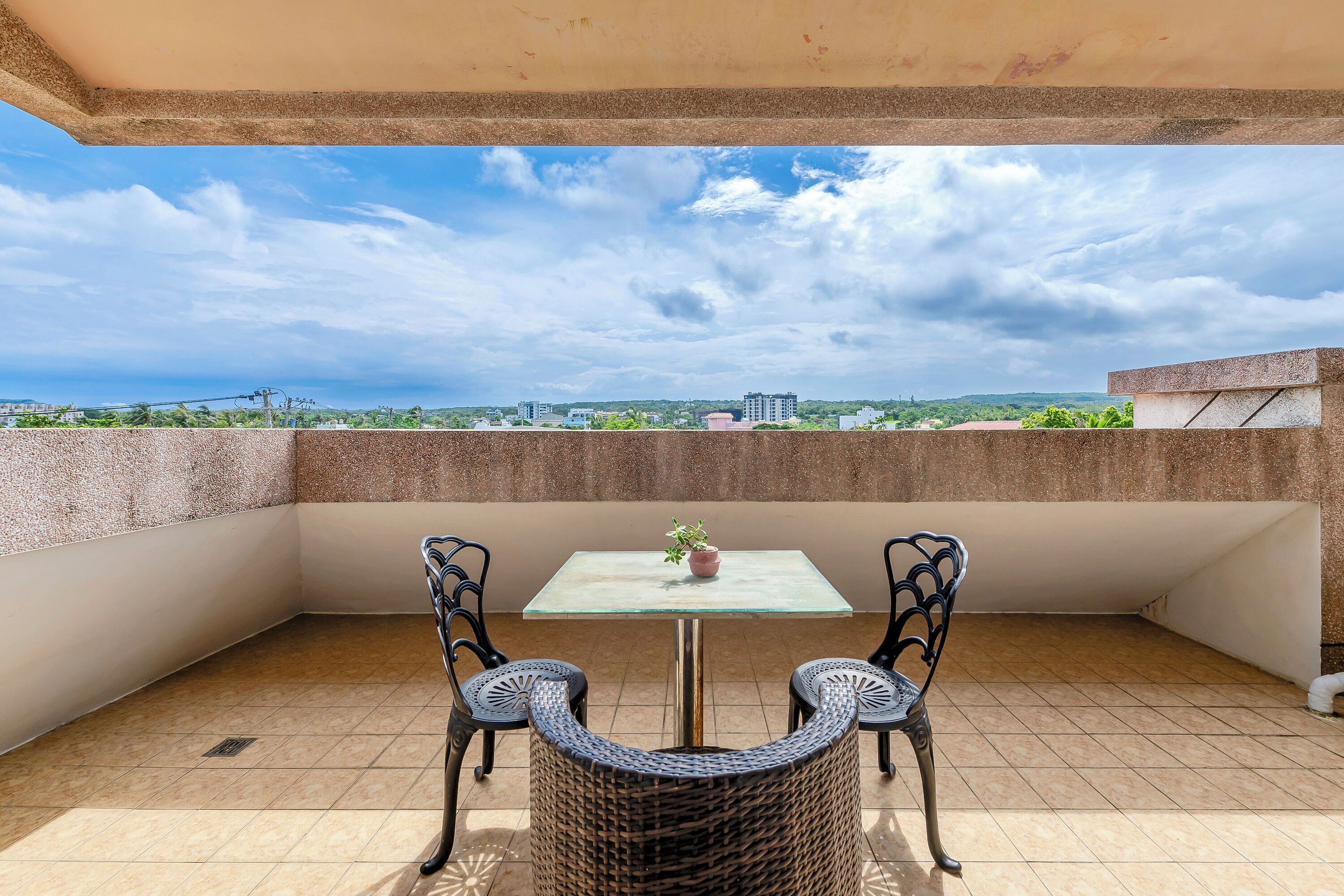 A balcony with a table and two chairs, a potted plant, and a view of distant buildings and greenery.