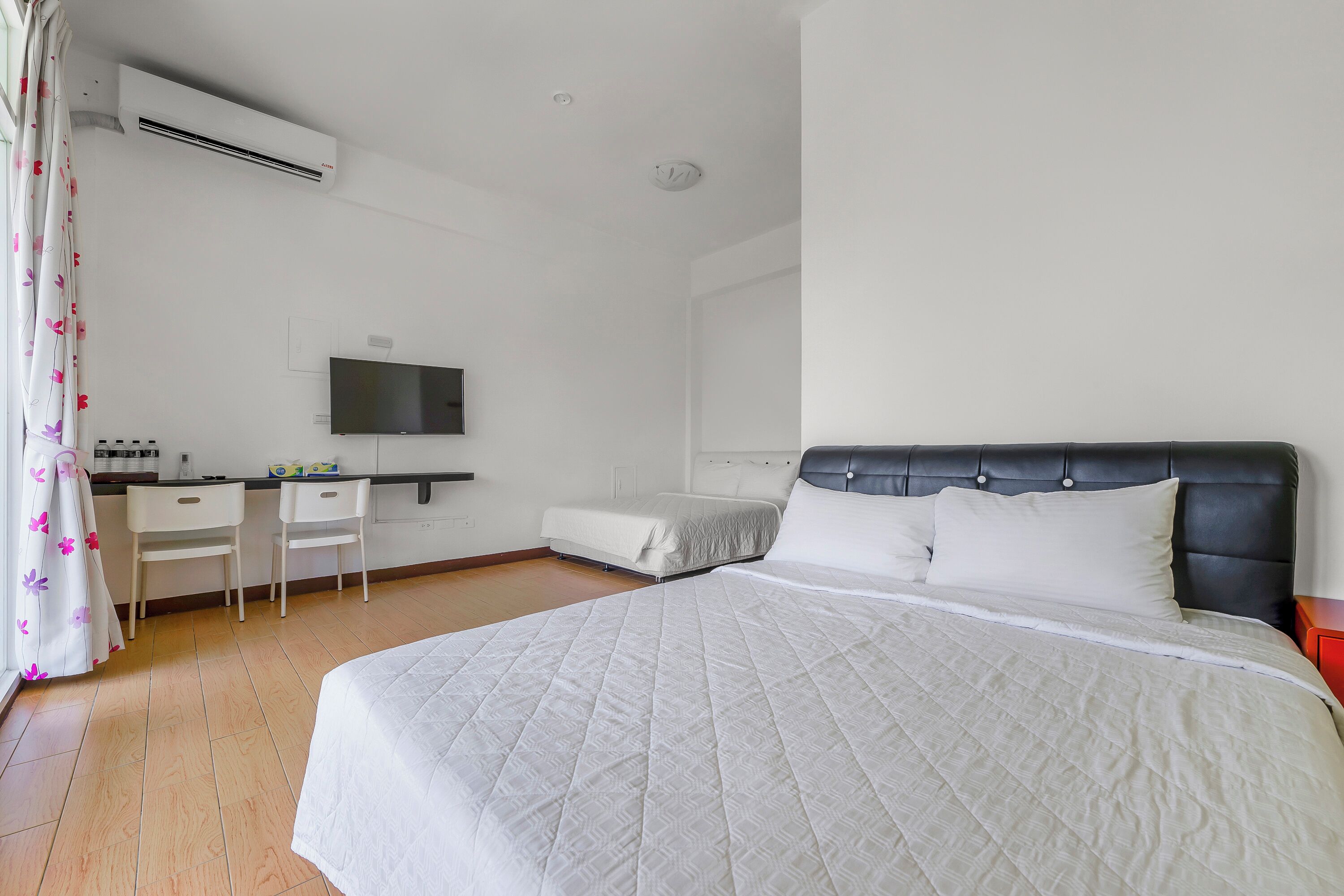 A hotel room with a bed, a desk, a chair, a TV, and an air conditioning unit.