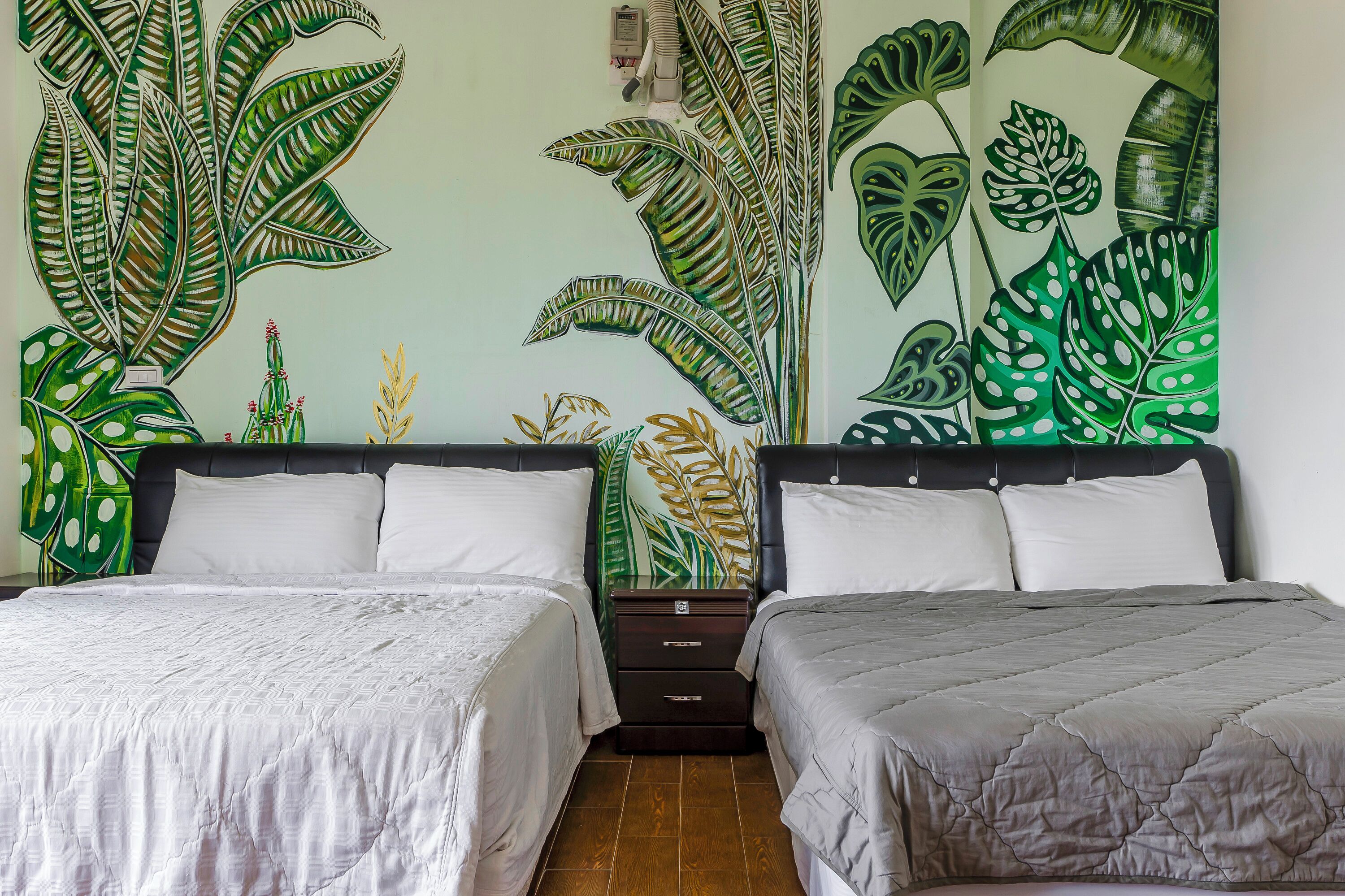 Une chambre avec deux lits, une table de chevet et une fresque murale représentant des feuilles tropicales.