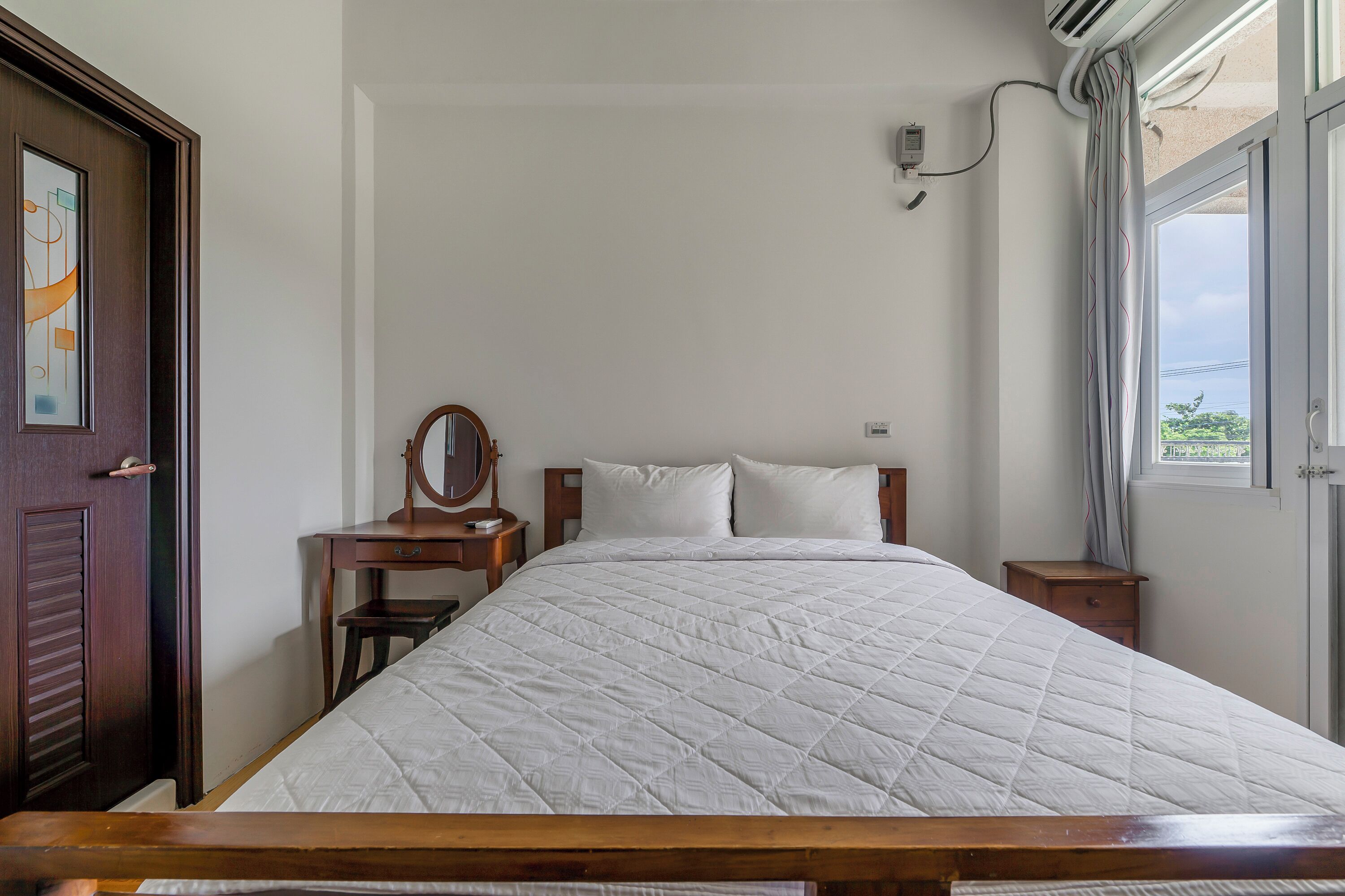 Een slaapkamer met een bed, een houten bureau, een spiegel en een raam met gordijnen.
