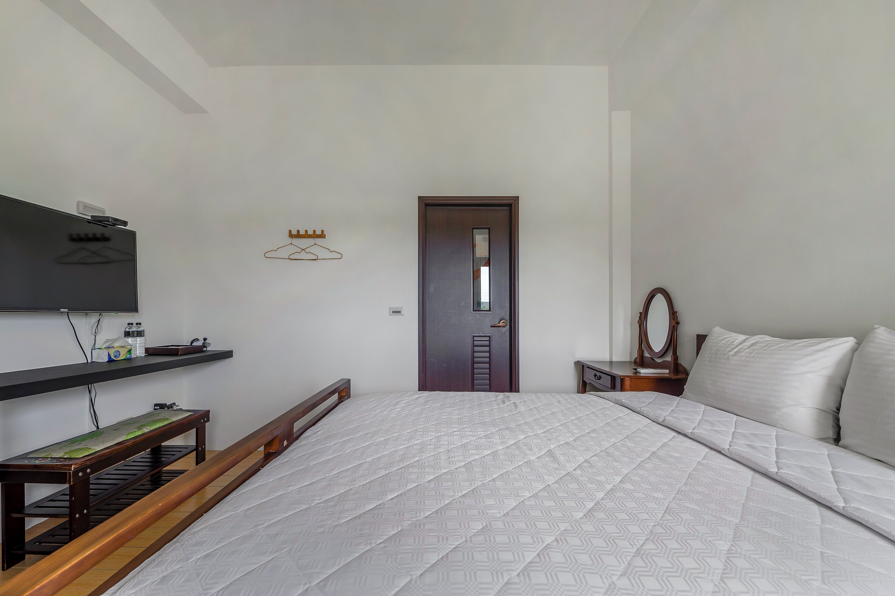 A bedroom with a bed, a wooden bench, a TV mounted on the wall, a door, and a small table with a mirror.