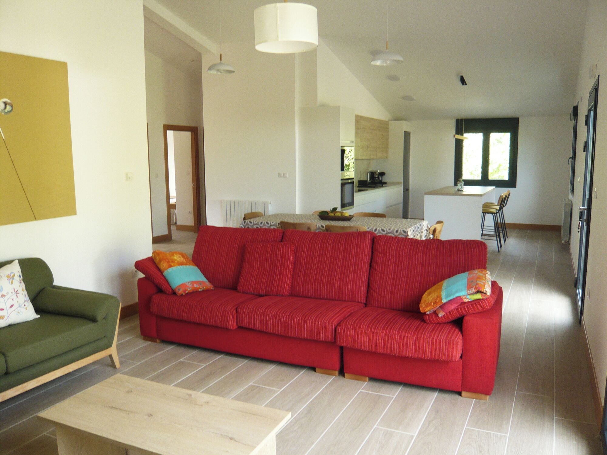 A spacious living area with a red sofa, a green armchair, and a wooden coffee table. A kitchen is visible in the background.