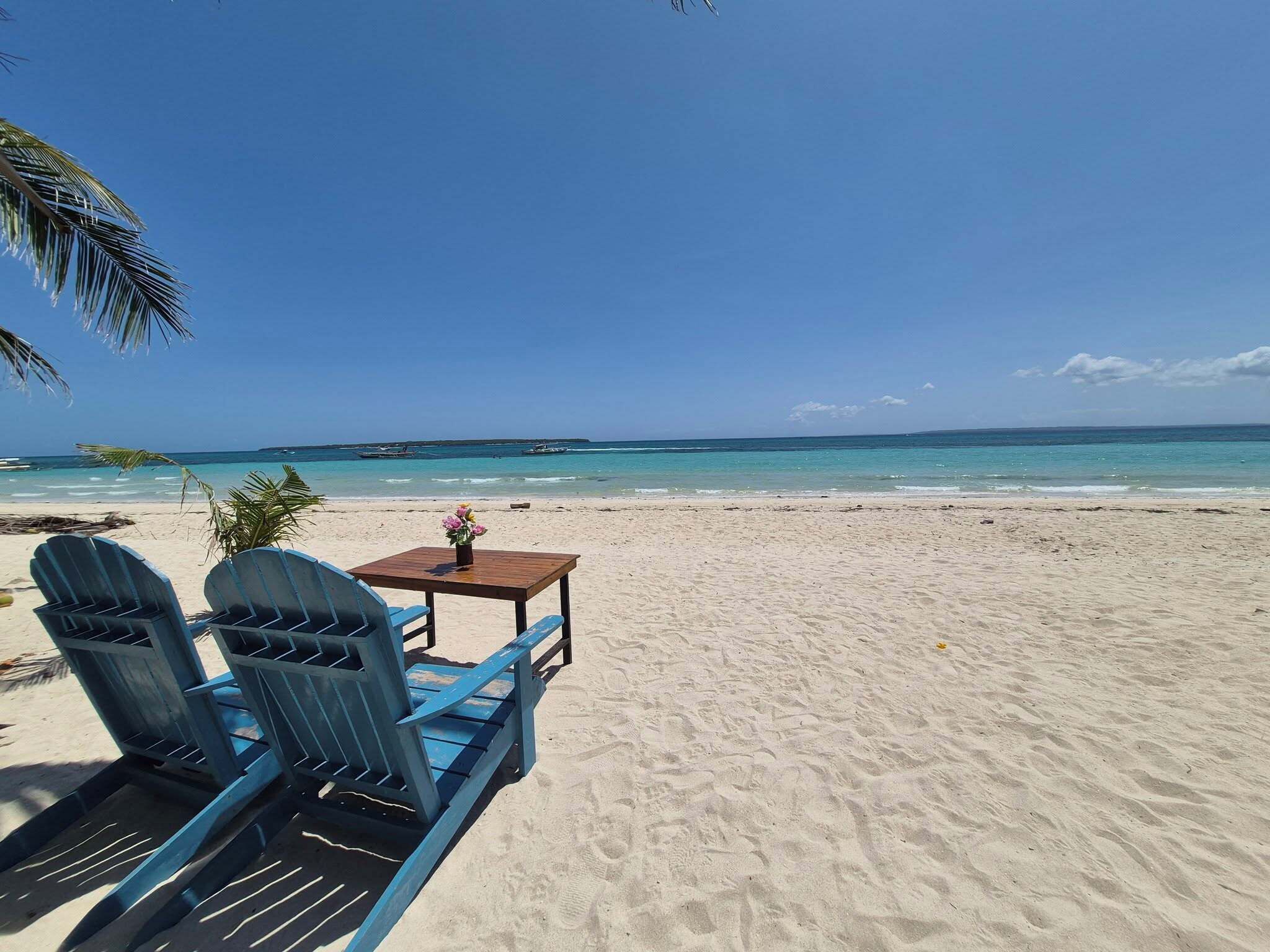 A beach with two blue chairs and a small table with a flower, under a clear blue sky.