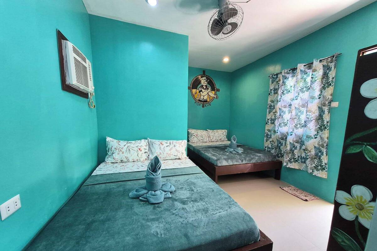 Una habitación con paredes color turquesa, una cama con manta gris, una mesita de noche, aire acondicionado de pared y un ventilador de techo, además de una cortina con estampado floral.