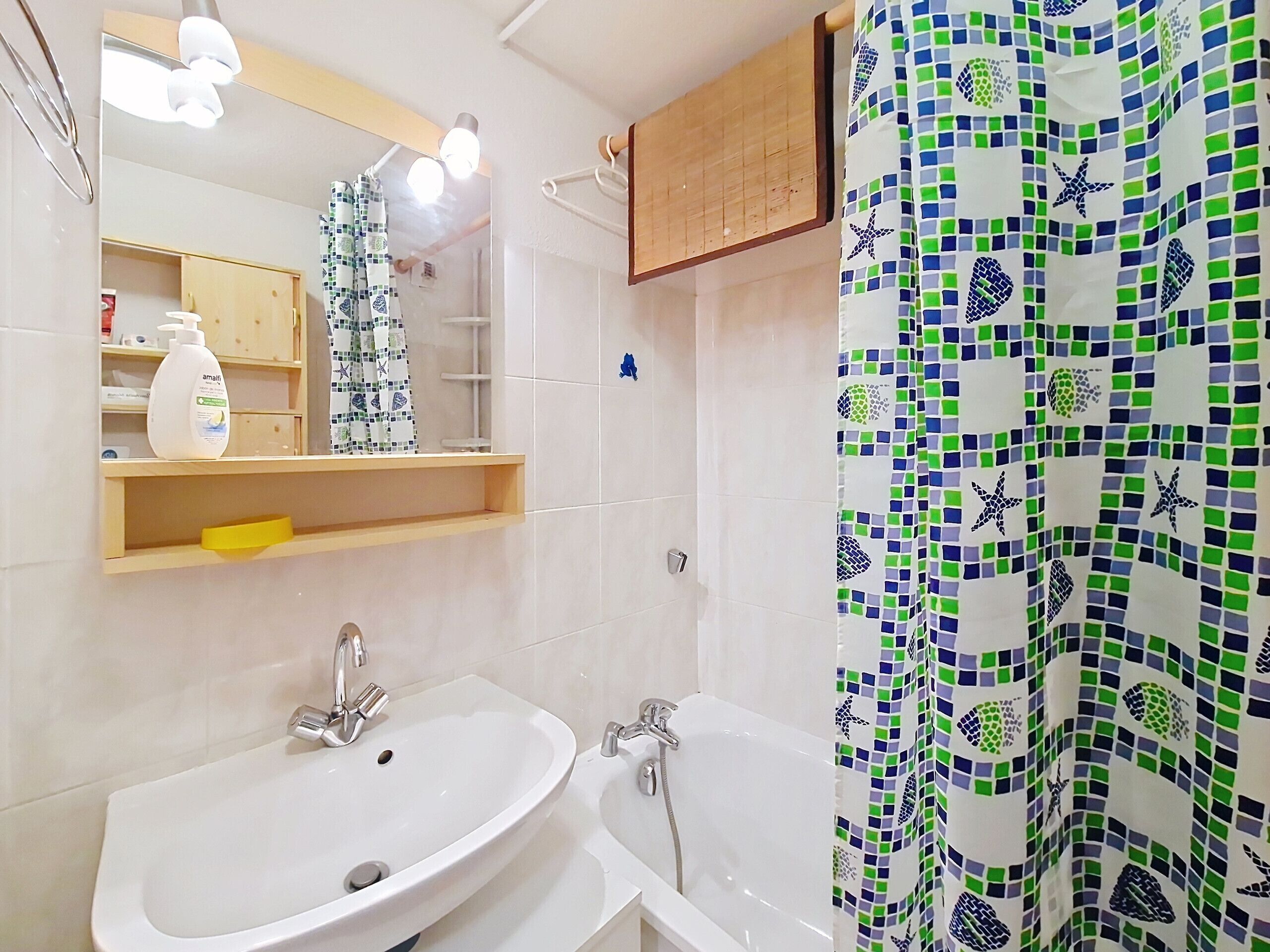 Baño con lavamanos, bañera y espejo. Se ve una repisa con artículos de aseo personal y una cortina de ducha con un diseño de mosaico.