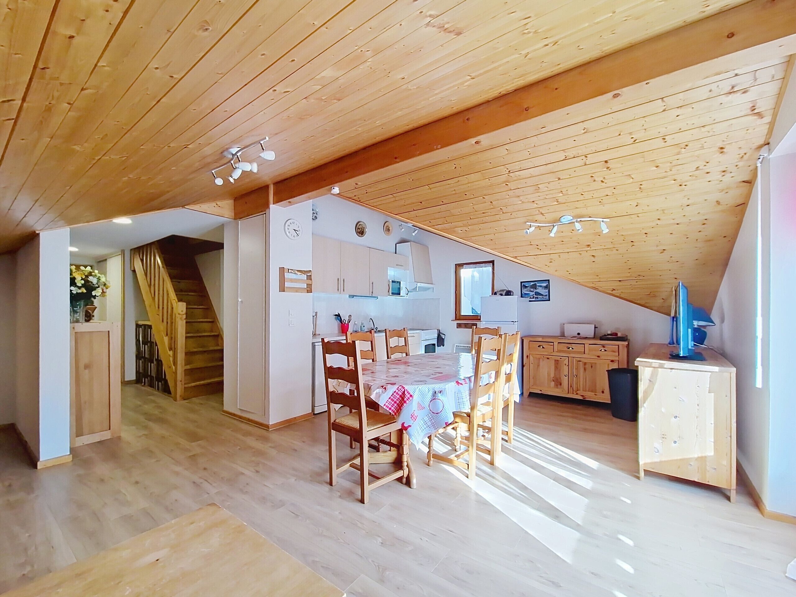 A spacious attic room with a wooden ceiling, a dining area with a table and chairs, a kitchen with cabinets and a refrigerator, and a small desk with a computer.