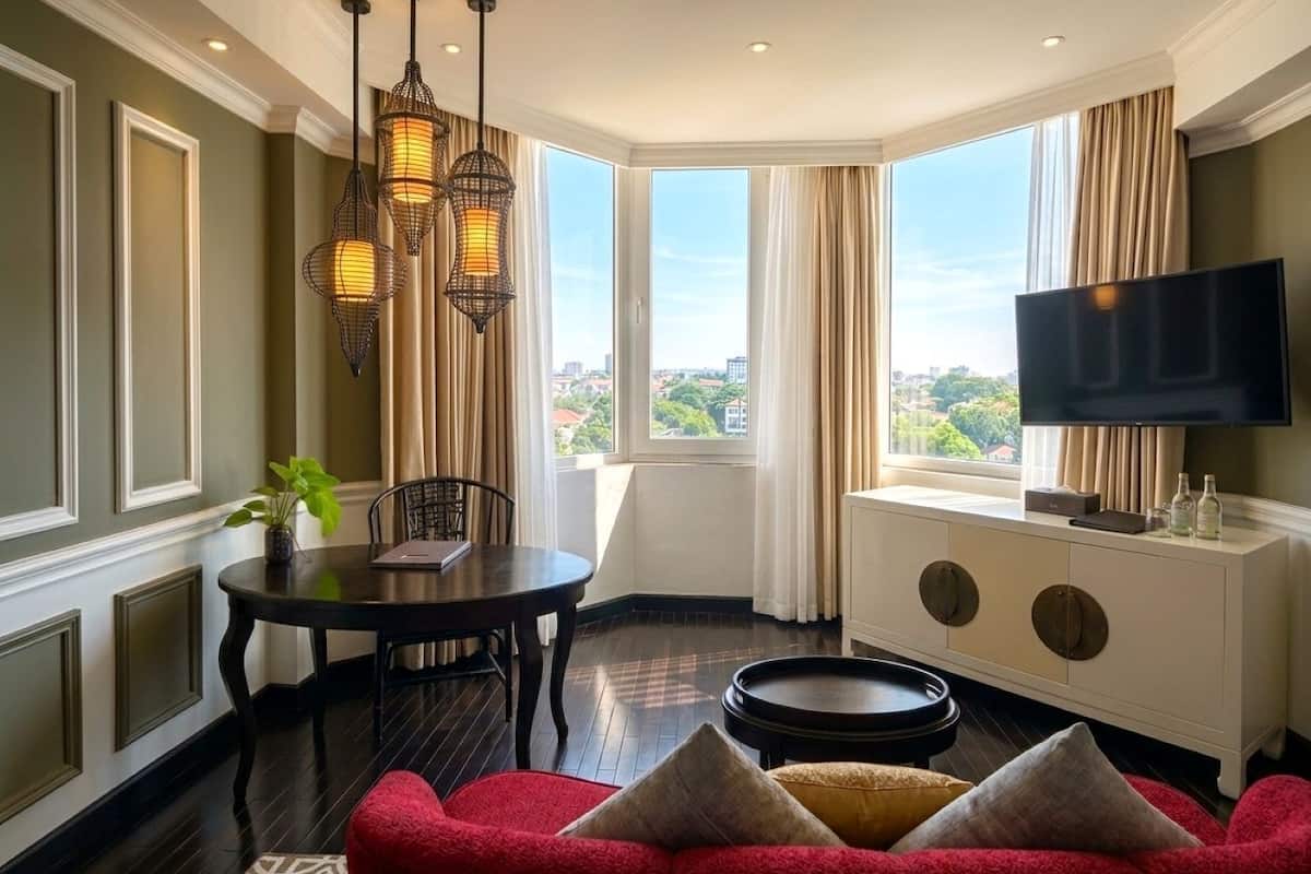 Habitación de hotel con mesa de comedor, sofá rojo, televisor y ventanas amplias con vista a la ciudad.