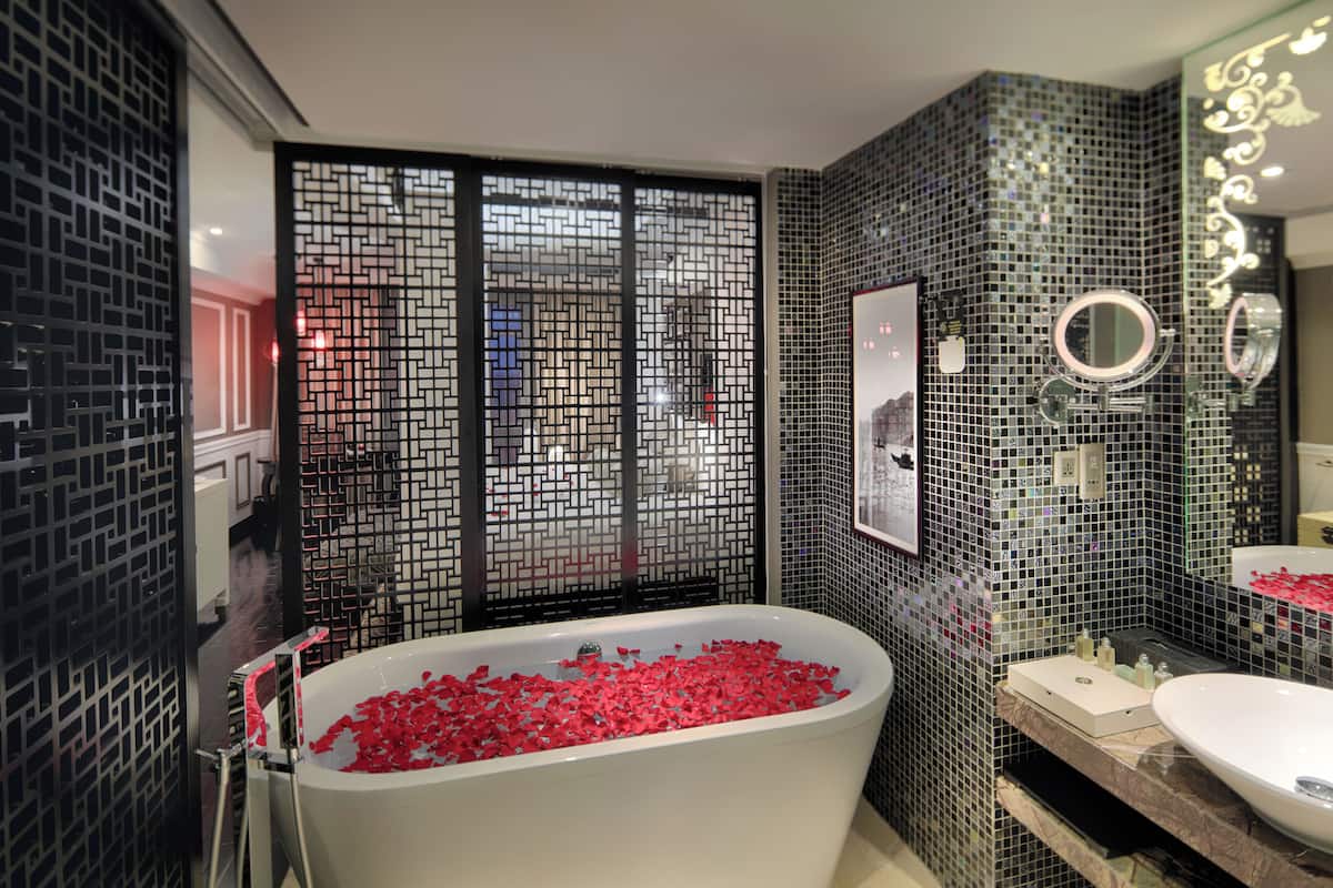 Un baño moderno con una bañera independiente llena de pétalos de rosa, un espejo grande y un panel decorativo en la pared.
