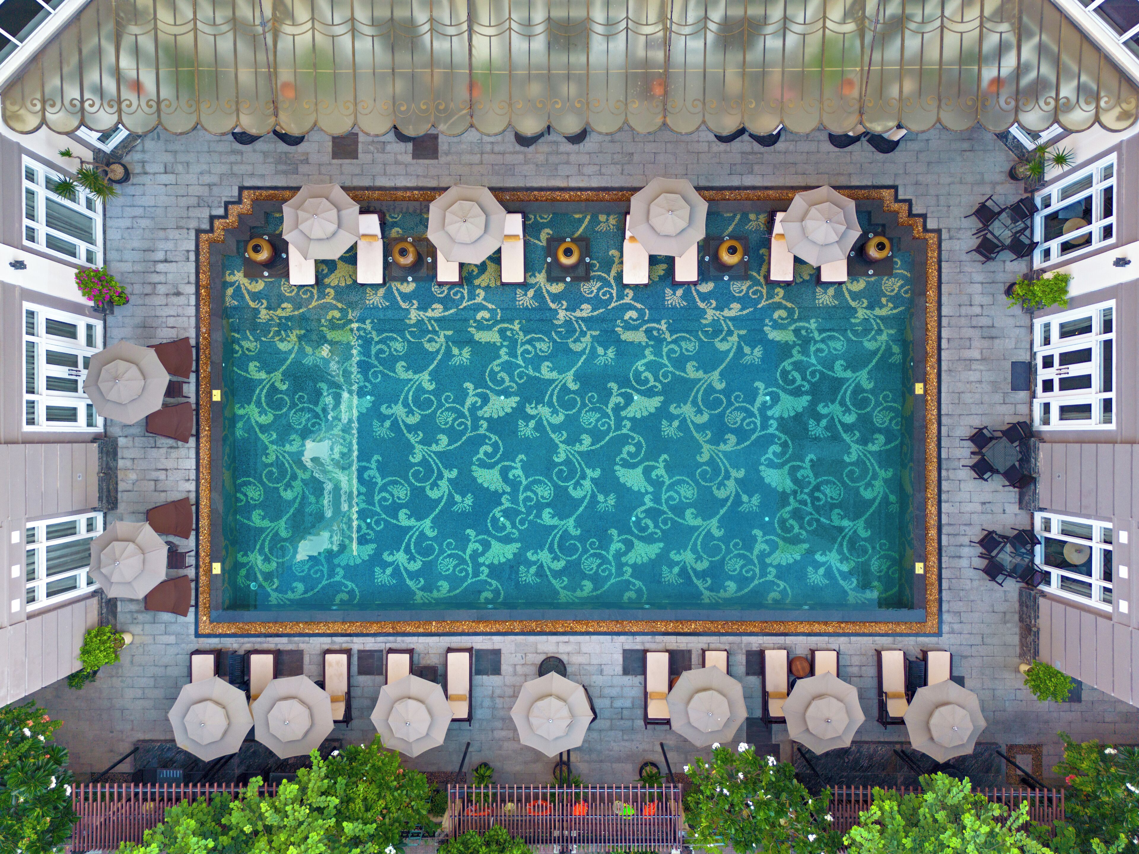 An aerial view of a rooftop pool area with a patterned floor, surrounded by lounge chairs and umbrellas, and bordered by a decorative railing.