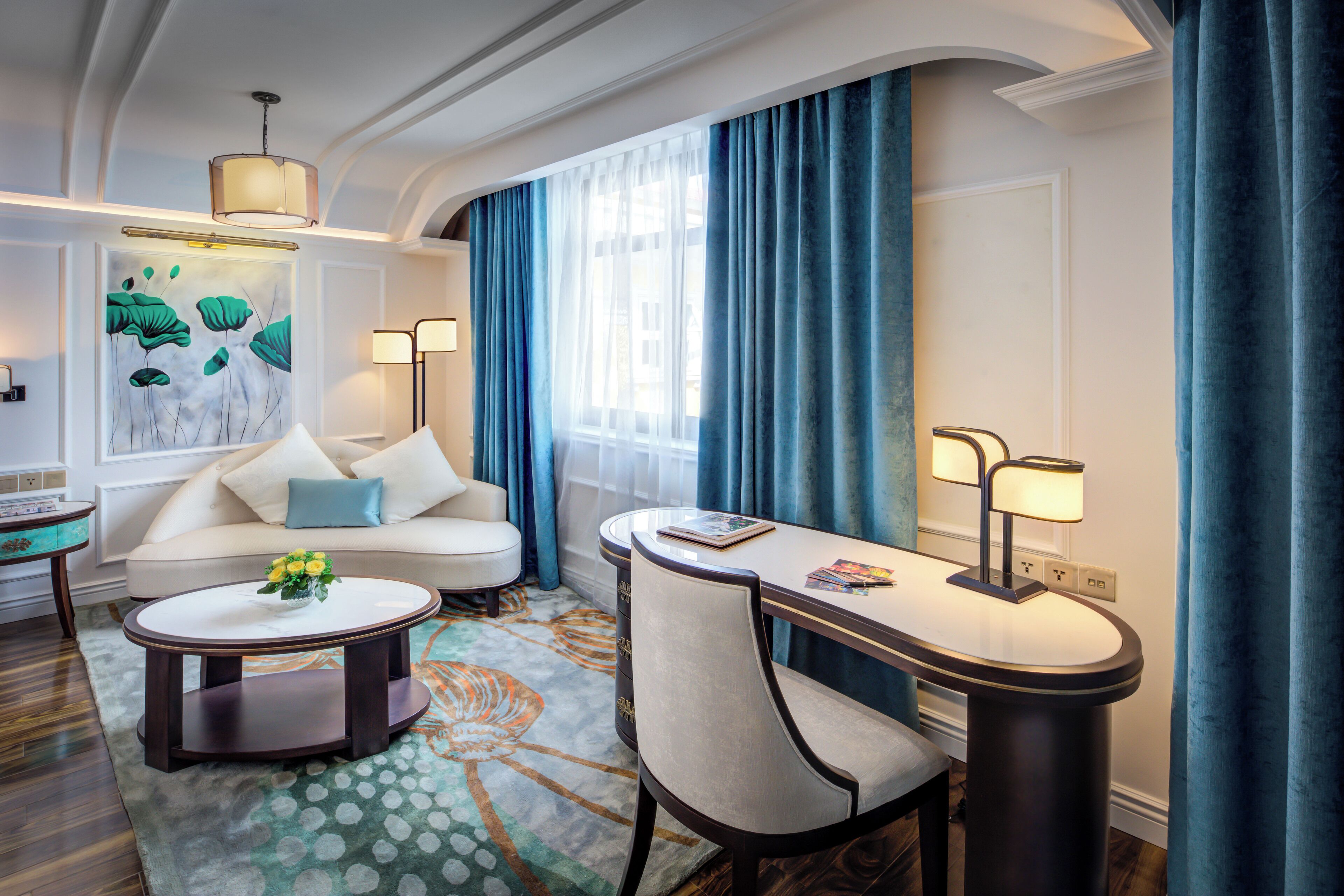 A modern hotel room with a sofa, a round coffee table, a dining table with chairs, a painting on the wall, and a large window with curtains.