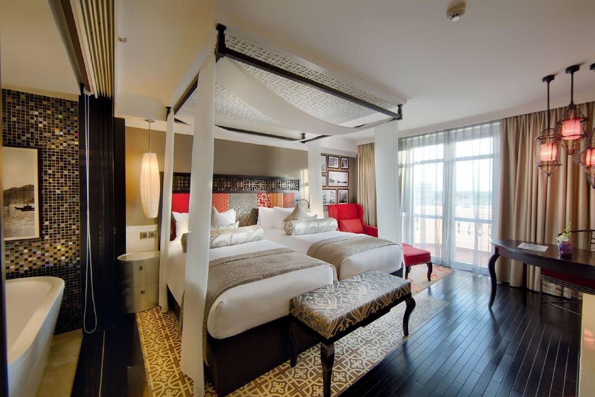 Habitación de hotel con cama con dosel, una zona de estar con una butaca roja, una mesa de comedor y un ventanal con cortinas.