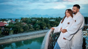 Una pareja con batas blancas junto a una barandilla, con vista a un río, edificios y follaje en el fondo.