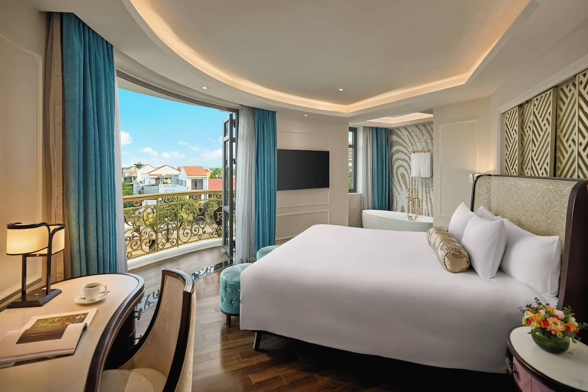 Habitación de hotel con una cama grande, un escritorio con lámpara, una silla y vistas al balcón exterior.
