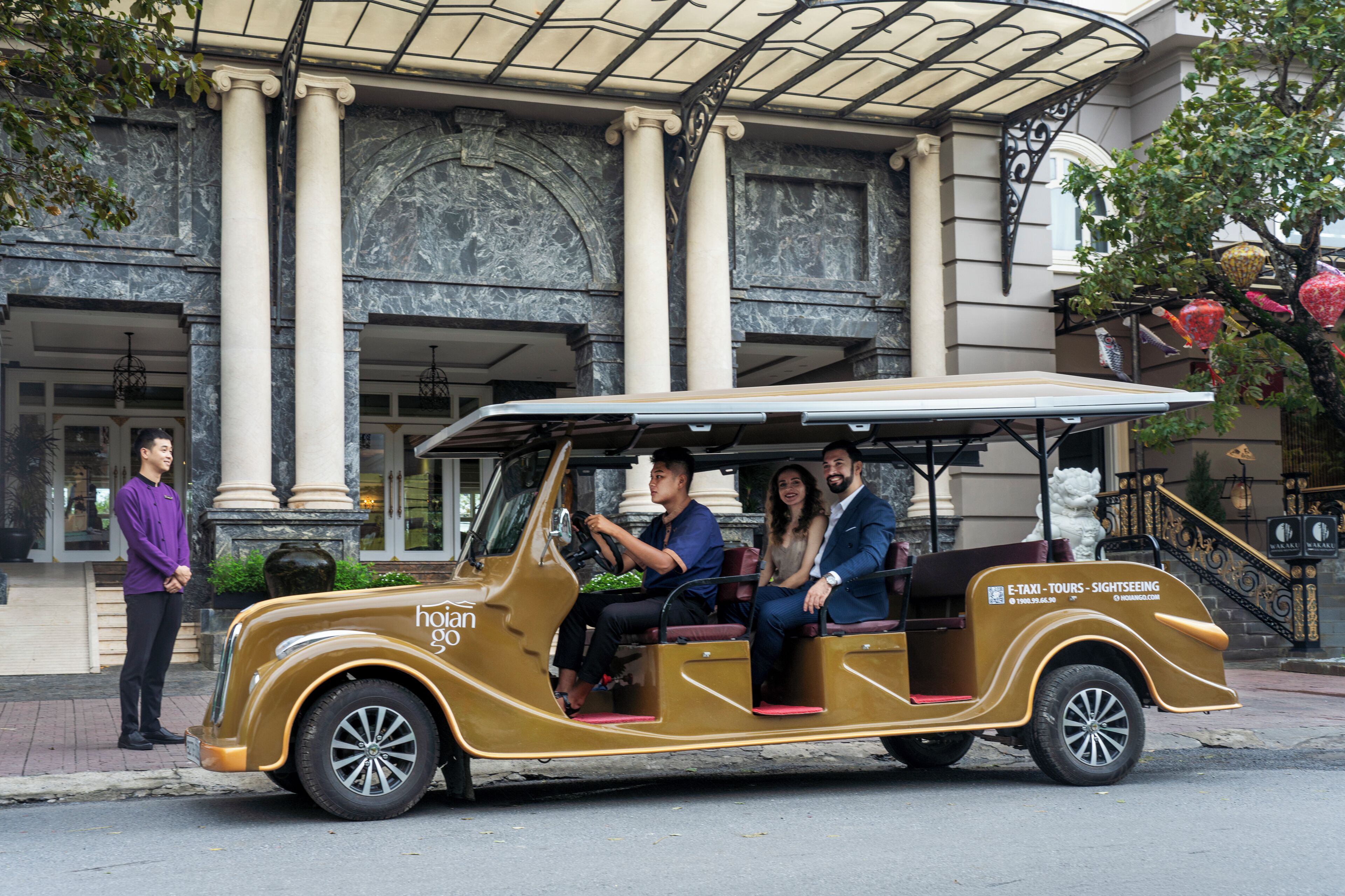 A vintage yellow car with passengers, parked in front of a building with columns and a statue.