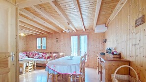 A wooden cabin interior with a dining table, chairs, a sofa, and a kitchen area. - Chalet Jet d'Eau JD20 NATURE & MOUNTAIN  8 Pers. (Châtel)
