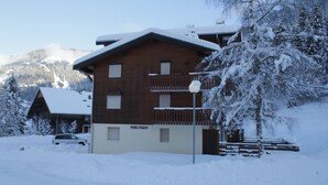 Ein zweistöckiges Holzhaus mit Balkon, umgeben von schneebedeckten Bäumen und einem Berg im Hintergrund.