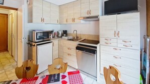 Eine kompakte Küche mit Holzausstattung, einer Mikrowelle, einer Kaffeemaschine und einem Geschirrspüler. Zwei Hocker aus Holz mit Herzförmigen Aussparungen sind vorhanden.