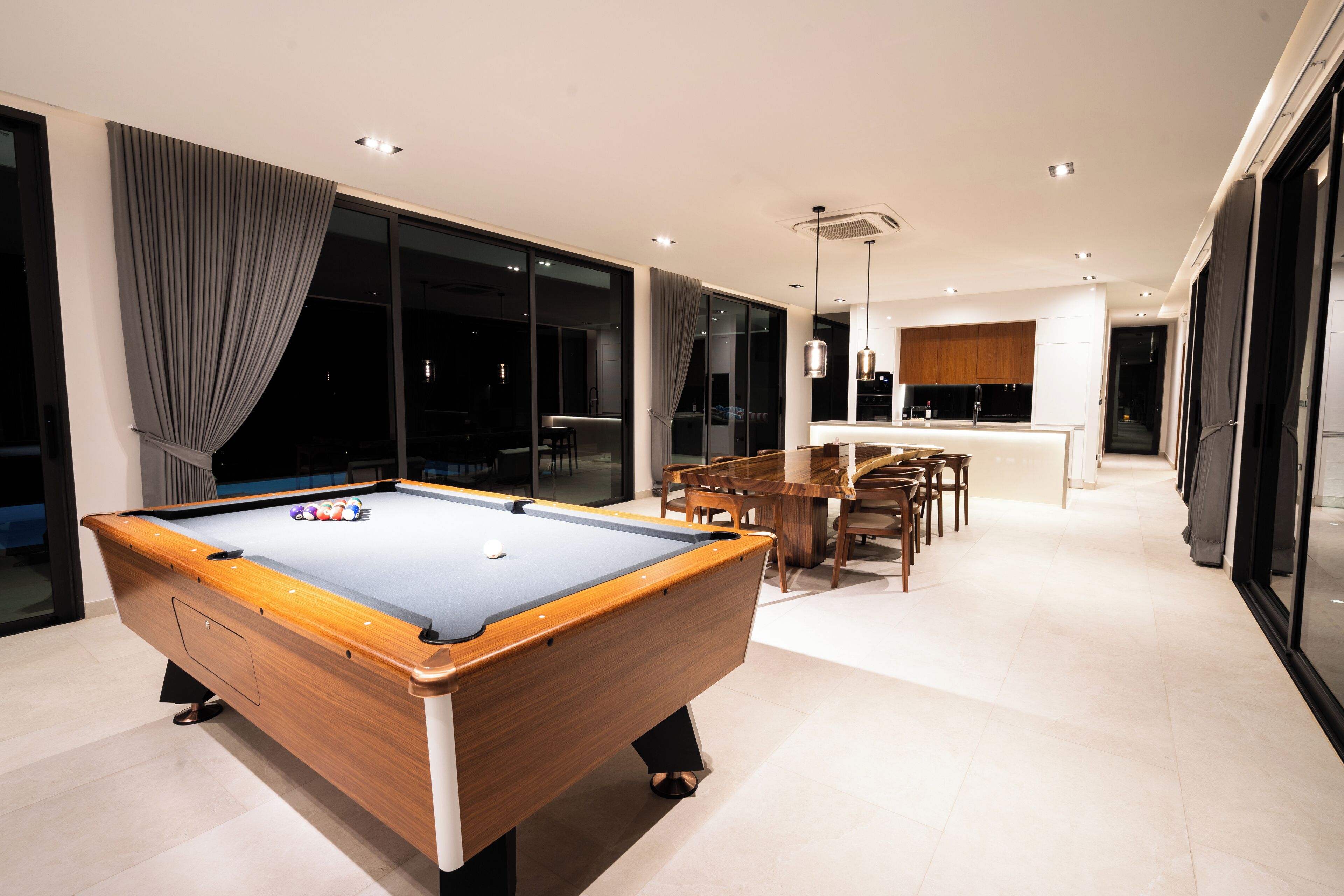 Una mesa de billar moderna en una habitación bien iluminada con ventanas amplias, un comedor y una cocina al fondo.