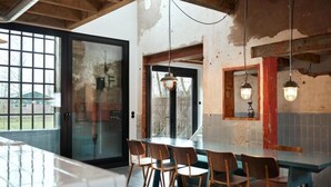 Een eethoek met een grote tafel en stoelen, een glazen deur naar buiten, en een rustiek interieur met onbehandelde bakstenen muren en houten balken.