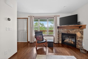 A living room with a stone fireplace, a television mounted above it, and a view of greenery outside.