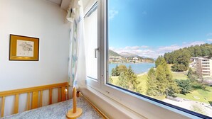 A room with a large window offering a view of a lake, buildings, and greenery. - Well-kept holiday apartament house in a prime location in St. (St. Moritz)